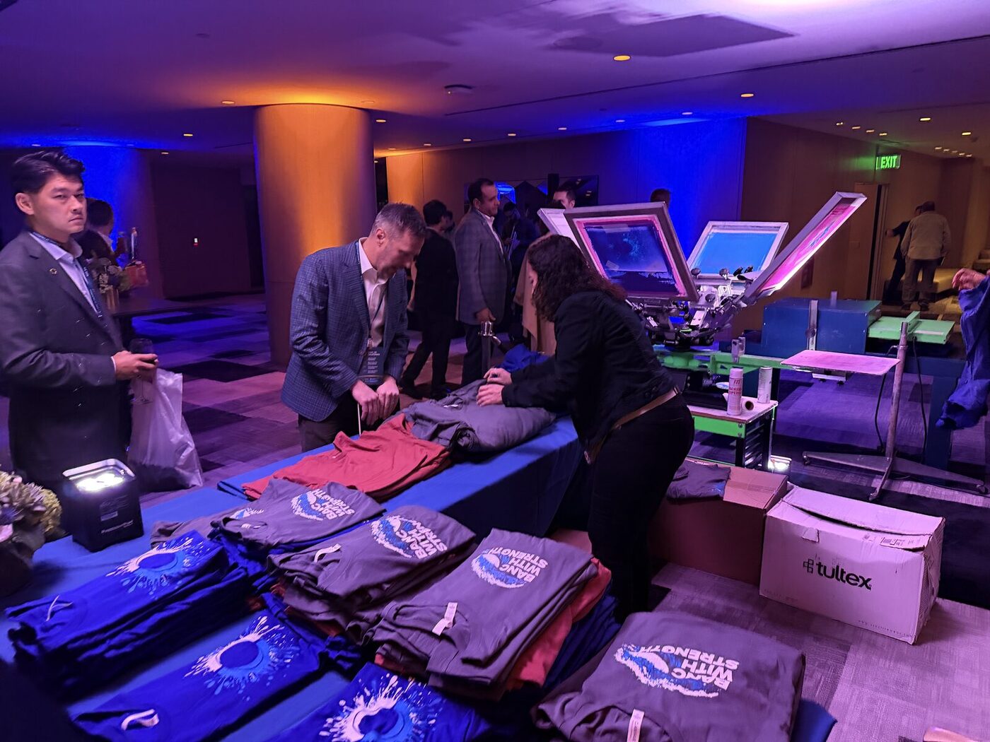 Guests browsing folded custom-printed shirts at a blue-lit evening conference reception